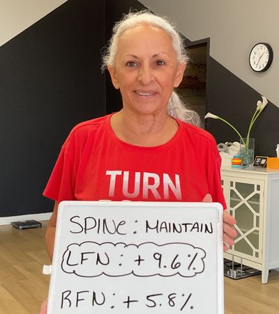 Ada, an OsteoStrong Austin member in her 60s, smiling and holding a whiteboard showing her DEXA bone-density gains: spine maintained, left femoral neck +9.6%, right femoral neck +5.8%